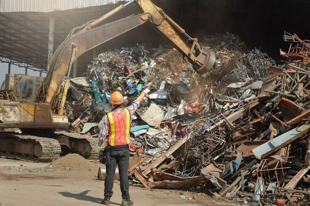 Worker directing excavator at scrap metal yard.